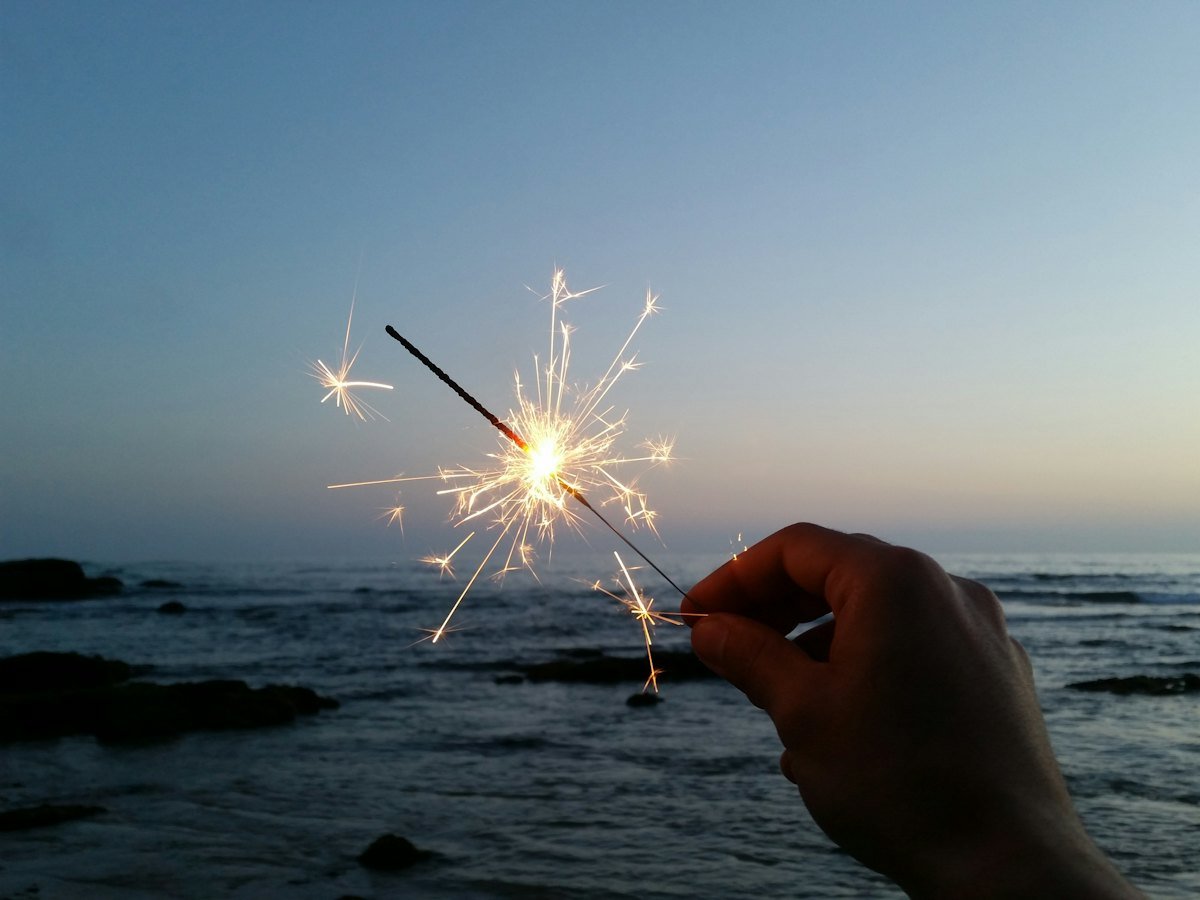 A hand holding a lit sparkler by the sea